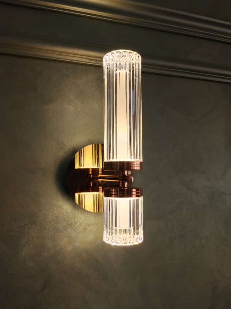 Modern kristal silindir tasarımlı çift yönlü LED duvar apliği, gold metal gövdesi ve şeffaf dekoratif cam detaylarıyla salon, oturma odası, yatak odası, antre, koridor, otel, restoran ve lüks iç mekân dekorasyonlarında kullanılan şık bir duvar aydınlatma modelidir. Yukarı ve aşağı yönlü LED ışık dağılımı sayesinde dekoratif duvar yüzeylerinde estetik bir ışık efekti oluşturarak modern yaşam alanlarına zarif ve dikkat çekici bir görünüm kazandırır. Açıklama Modern Kristal Silindir LED Aplik, yukarı ve aşağı yönlü çift taraflı aydınlatma sunan zarif tasarımıyla dekoratif duvar aydınlatmalarında öne çıkan özel modellerden biridir. Kristal desenli silindirik difüzörleri ve metal gövdesi, ışığın daha şık ve homojen yayılmasını sağlayarak yaşam alanlarına estetik bir atmosfer kazandırır. Salon, TV ünitesi, yatak odası, antre, koridor, otel, restoran, kafe ve özel projelerde tercih edilen bu model; lambiri, PVC mermer panel, dekoratif çıta ve modern duvar kaplama uygulamalarıyla kusursuz bir uyum oluşturur. Aynı modelin metal yüzey dokusu başta gold ve gümüş olmak üzere iki ana renk seçeneğiyle sunulmaktadır. Bazı modellerde ayrıca siyah, füme ve bronz renk alternatifleri de bulunmaktadır. DecoRoyal, dekoratif aydınlatma ve duvar tasarım ürünlerinde kaliteli ürün, güvenilir hizmet ve Türkiye geneline hızlı sevkiyat desteği sunmaktadır. Ayrıntılı bilgi ve sipariş için www.decoroyal.com.tr adresini ziyaret edebilir veya 0545 825 44 66 numaralı WhatsApp hattımızdan bizimle iletişime geçebilirsiniz. Aplik modelleri, Avize modelleri, Dekoratif aydınlatma, LED aplik modelleri, Kristal aplik, Duvar apliği, Modern duvar lambası, Çift yönlü aplik, Ev aydınlatma, İç mekân aydınlatma, Wall light models, Chandelier models, Decorative lighting, Crystal wall light, Home lighting, Wandleuchten Modelle, Kronleuchter Modelle, Dekorative Beleuchtung, Kristall Wandleuchte, Wohnraumbeleuchtung, Modèles d'appliques murales, Modèles de lustres, Éclairage décoratif, Applique murale en cristal, Éclairage intérieur, Modelli di applique, Modelli di lampadari, Illuminazione decorativa, Applique in cristallo, Illuminazione per la casa, Модели настенных светильников, Модели люстр, Декоративное освещение, Хрустальный настенный светильник, Домашнее освещение, موديلات أباليك, موديلات ثريات, إضاءة ديكورية, أبليك كريستال, إضاءة منزلية.